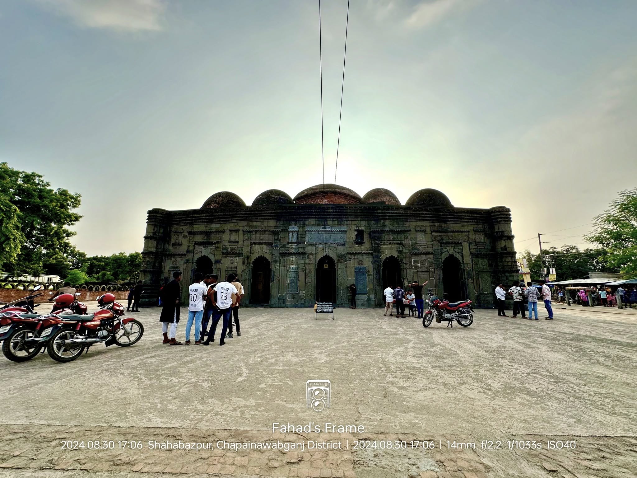 A wide view of an ancient six-domed mosque with people and motorbikes in the foreground.