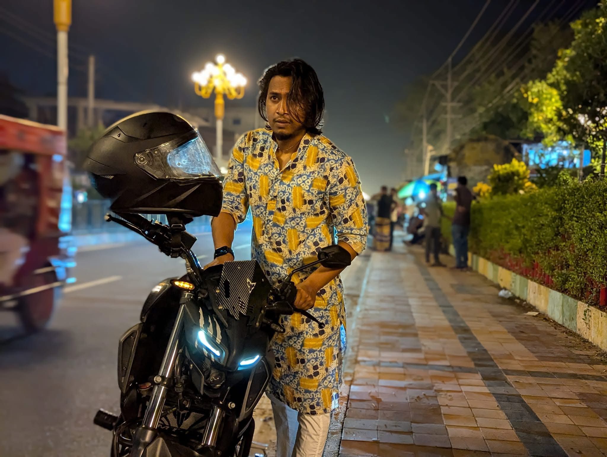 A man standing next to a motorcycle at night under warm street lights.